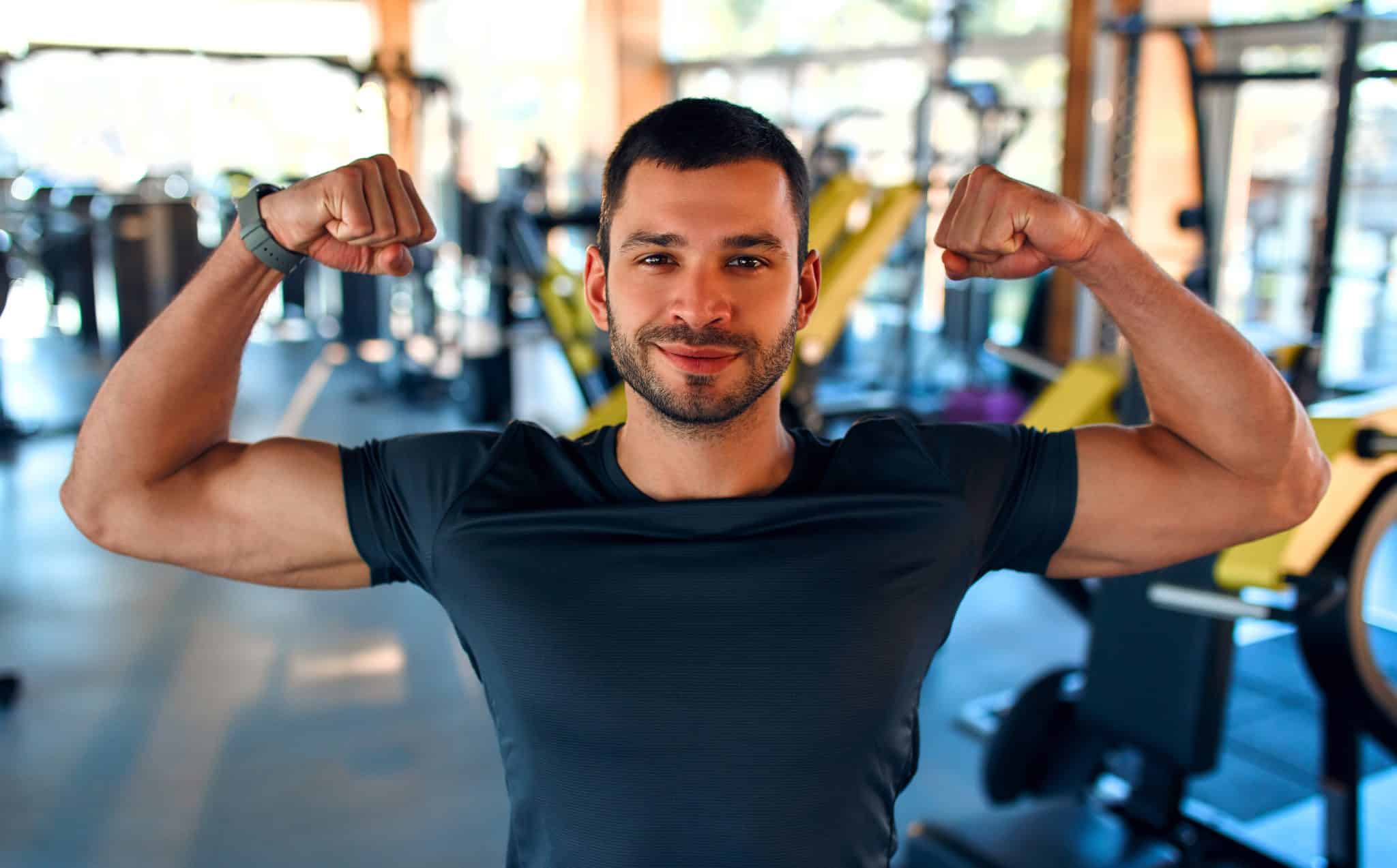 A man flexing his arm muscles and smiling inside a gym.