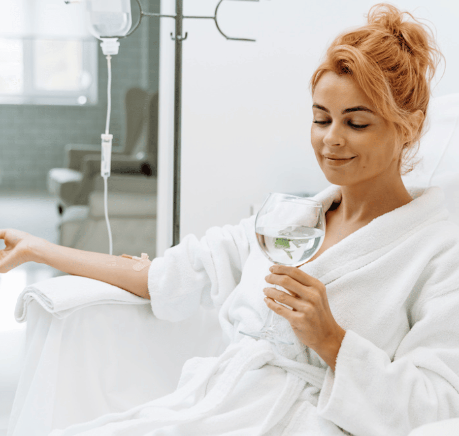 A charming woman in white bathrobe sitting in armchair and receiving IV infusion. She is holding glass of lemon beverage and smiling.