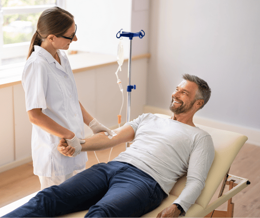 Man lying down and smiling attached to an IV bag and a nurse monitoring them.