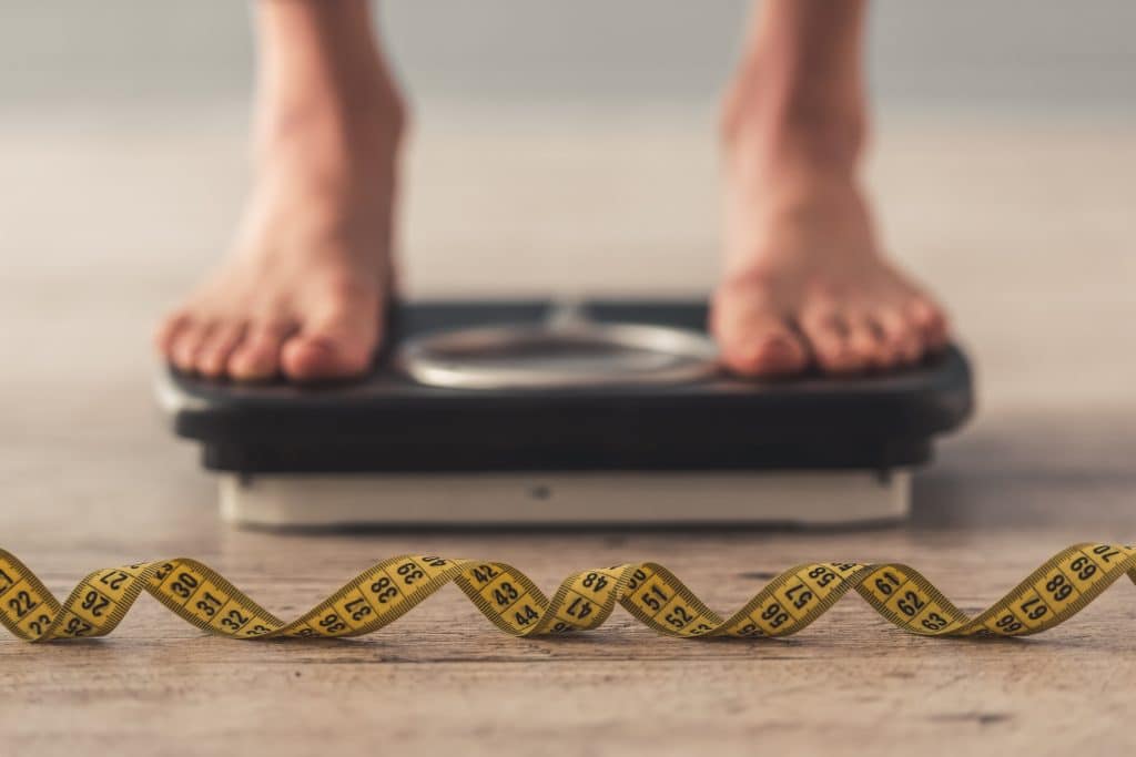 Cropped image of woman feet standing on weigh scales, on gray background. A tape measure in the foreground