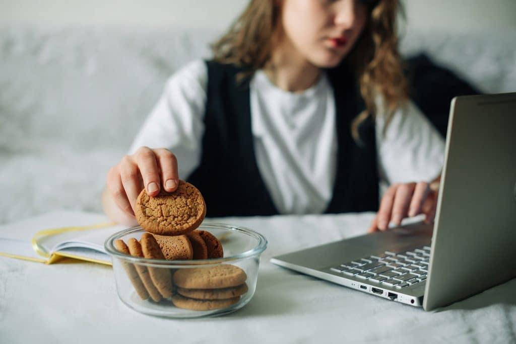 Unconscious eating. Bad habits. Selective focus of hand of concentrated female working on laptop at the kitchen table, taking unintentionally and automatically cookies from a transparent glass jar.