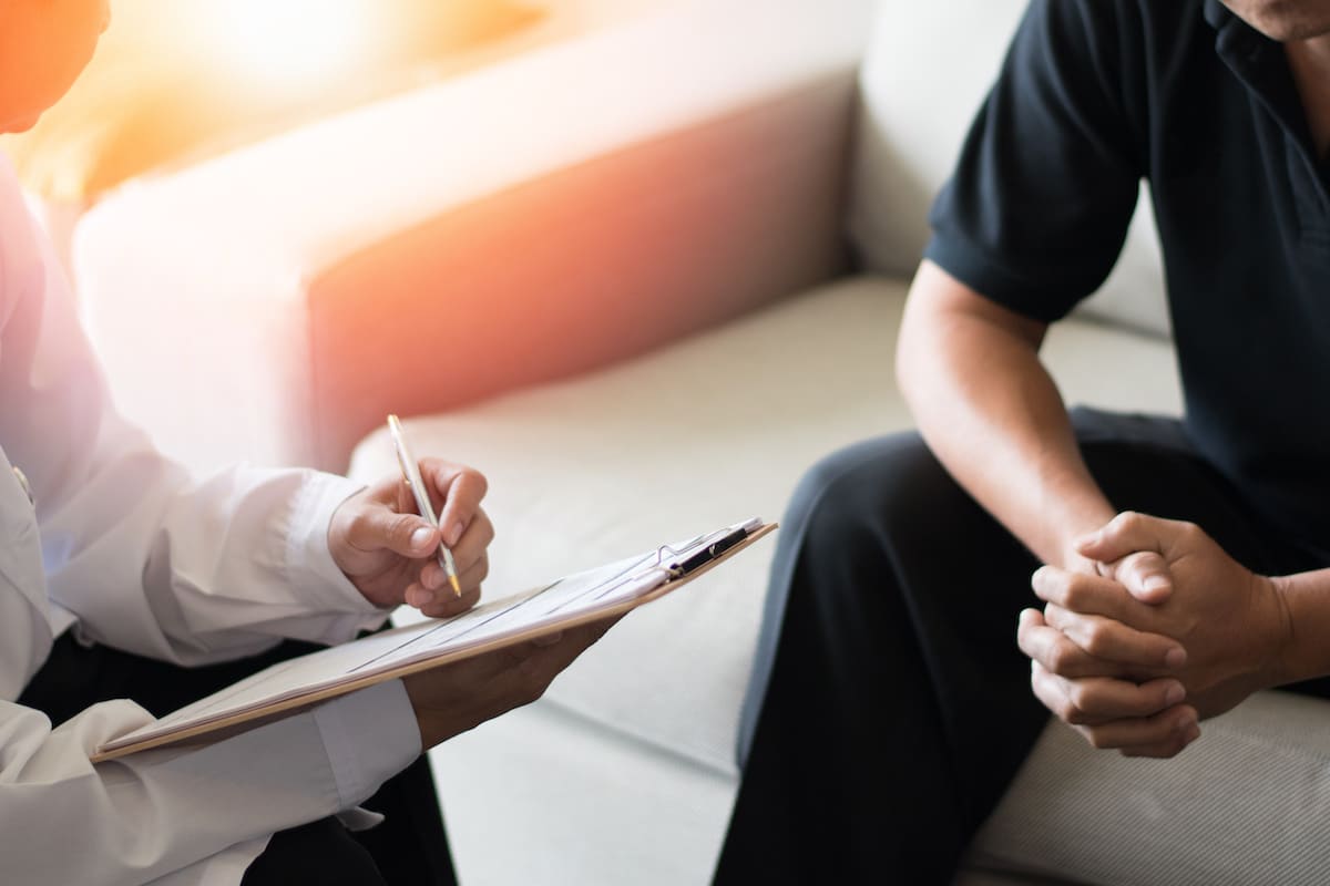 A doctor consulting with a patient, taking notes while the patient sits opposite, hands clasped.