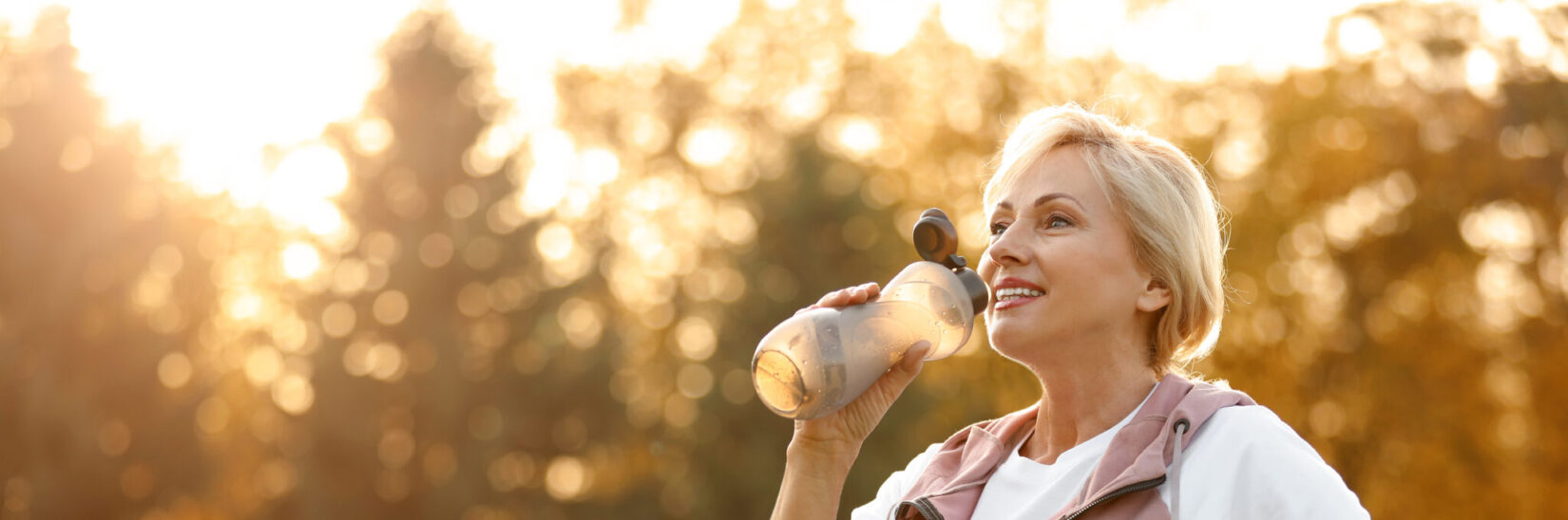 A healthy middle-aged woman drinks water outdoors, wearing workout clothes.