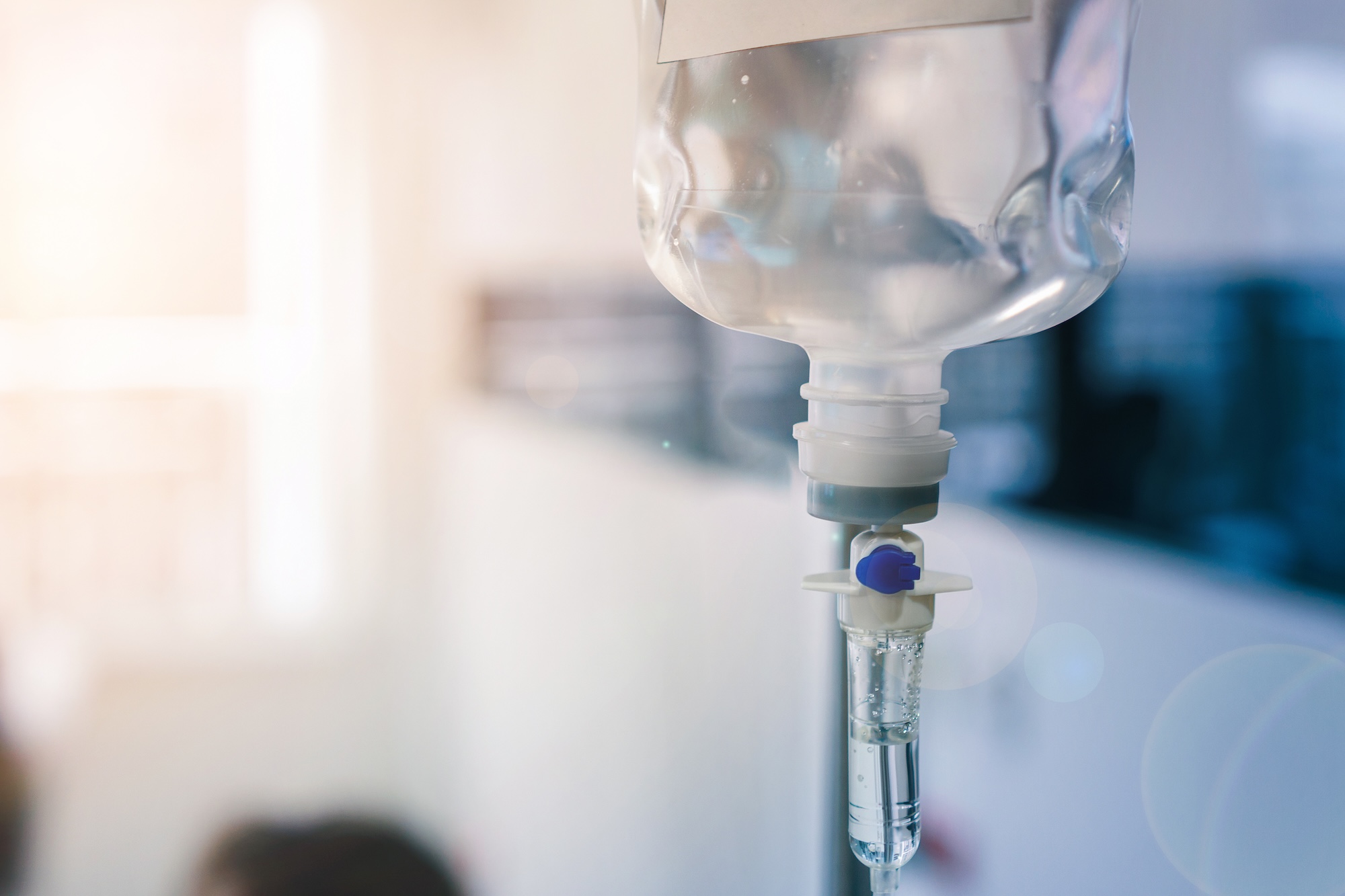 Close-up of an intravenous (IV) fluid bag with clear liquid inside, connected to a drip chamber and tubing, in a brightly lit medical setting.
