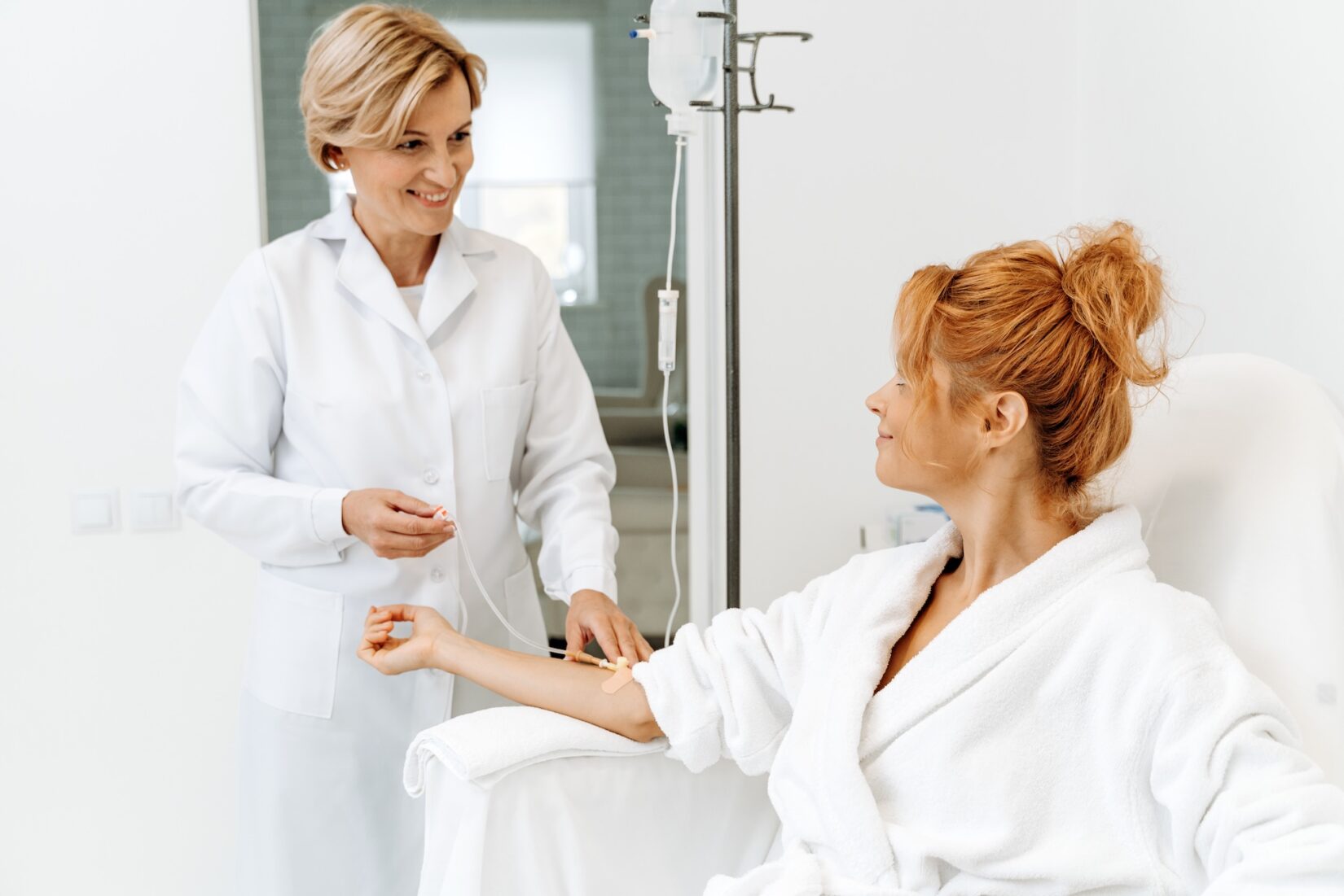 A healthcare professional in a white coat is preparing an IV drip for a woman seated in a chair.