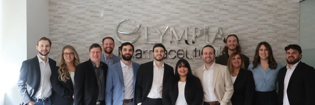 A wide shot of multiple members of Olympia's Business Development Team. Members from the Commercial and Patient Sales teams can be seen smiling together, in professional attire, in front of a silver logo that reads "Olympia Pharmaceuticals."
