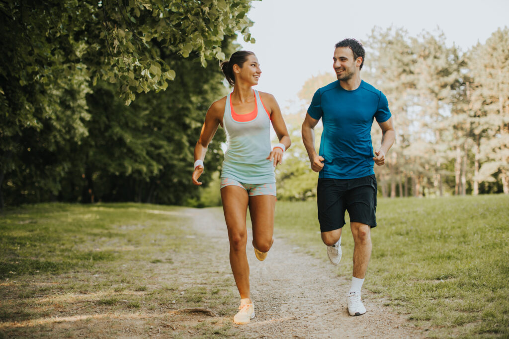 Athletic man and woman jogging outdoors on a tree-lined path, smiling at each other during their workout.