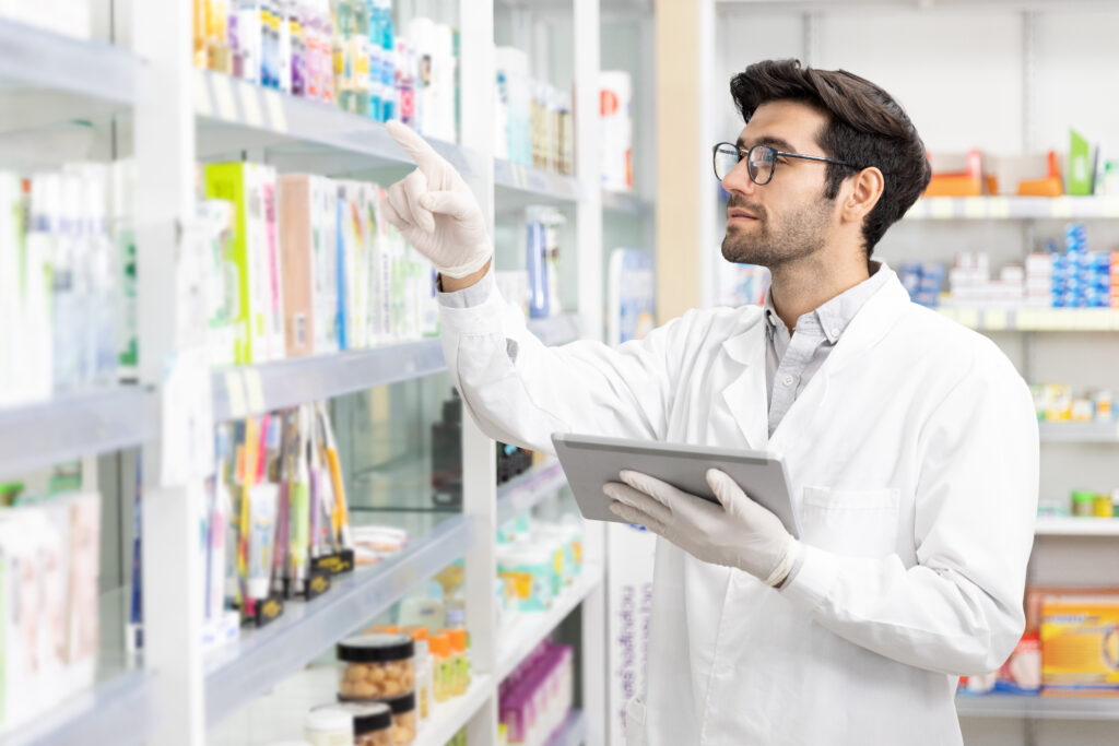 Male pharmacist wearing a lab coat and gloves reviews inventory on a pharmacy shelf while holding a tablet.