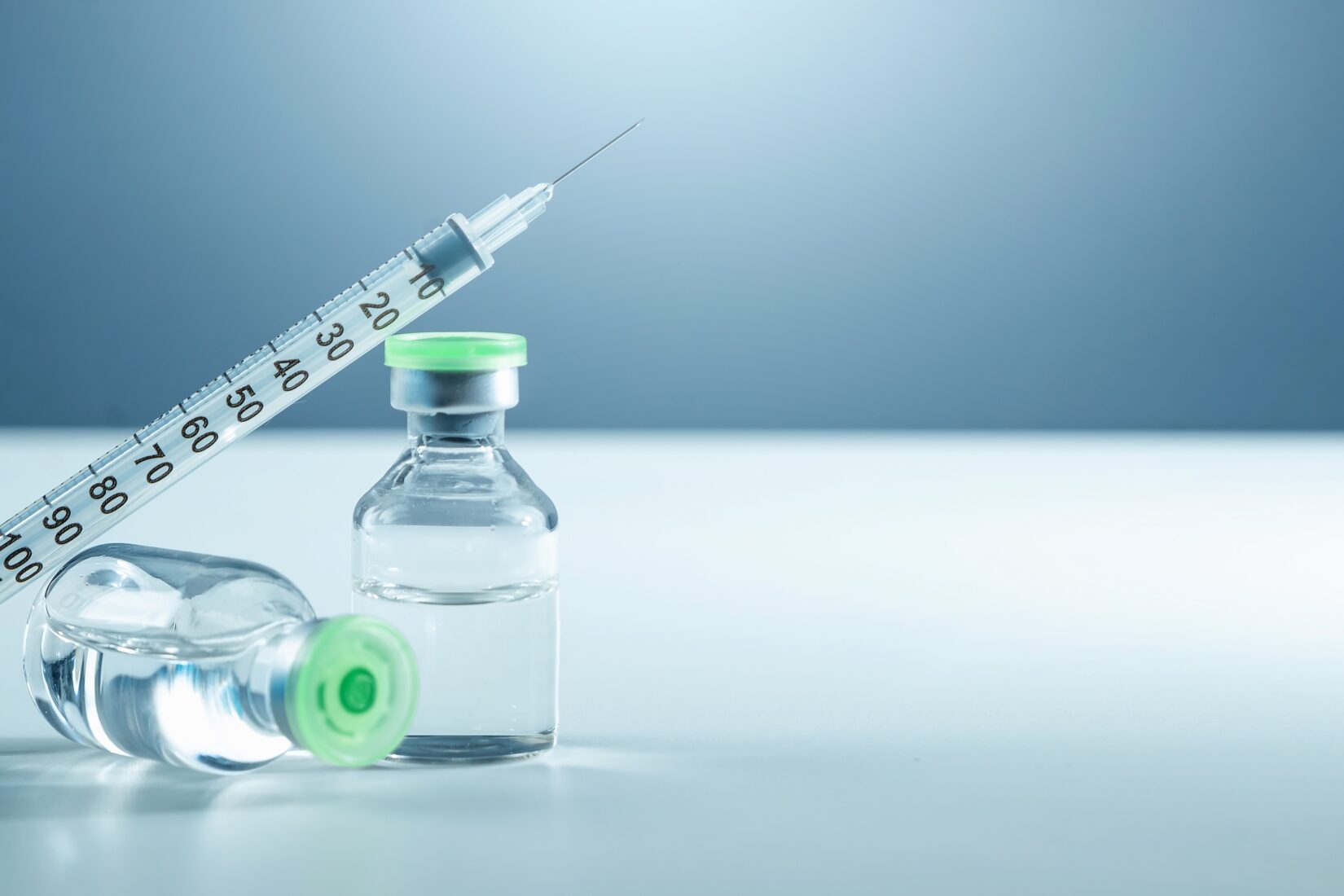 Syringe resting on two clear vials filled with liquid medication on a white surface with a blue background.