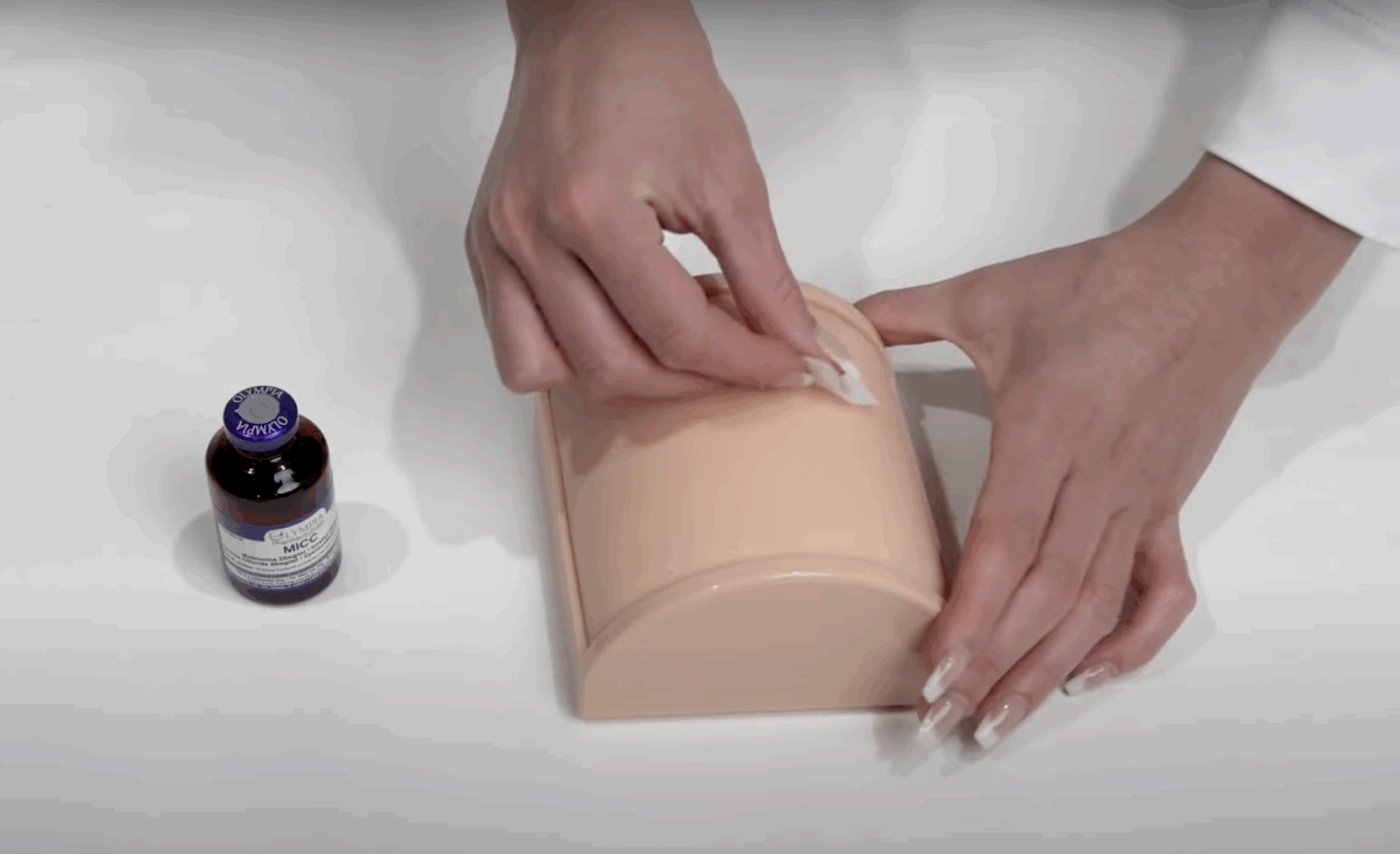 Hands sanitizing a practice injection pad with an alcohol swab next to a vial of Olympia MICC medication.