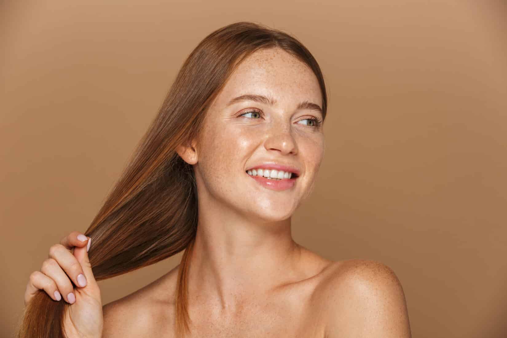 Woman with long straight hair and freckles smiling and holding her hair against a neutral background.