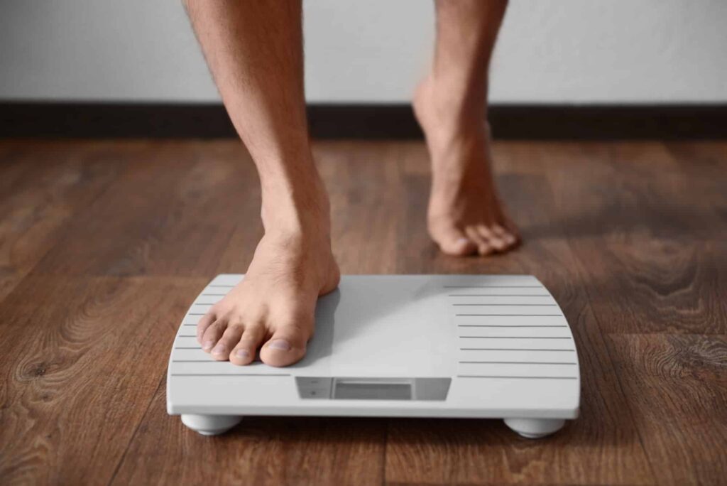 A person standing with one foot on a digital bathroom scale placed on a wooden floor.