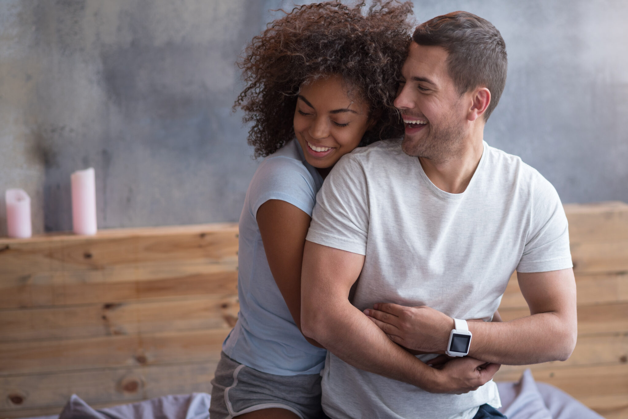 Happy couple sitting together on a bed, with the woman hugging the man from behind and both smiling warmly in a relaxed, intimate moment.