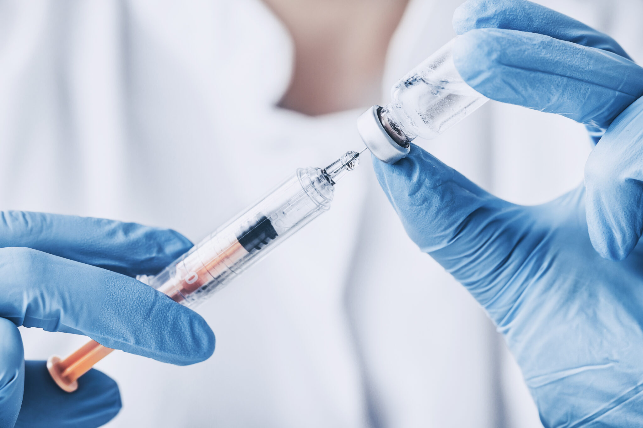 Close-up of gloved hands drawing liquid from a vial into a syringe, preparing an injection.