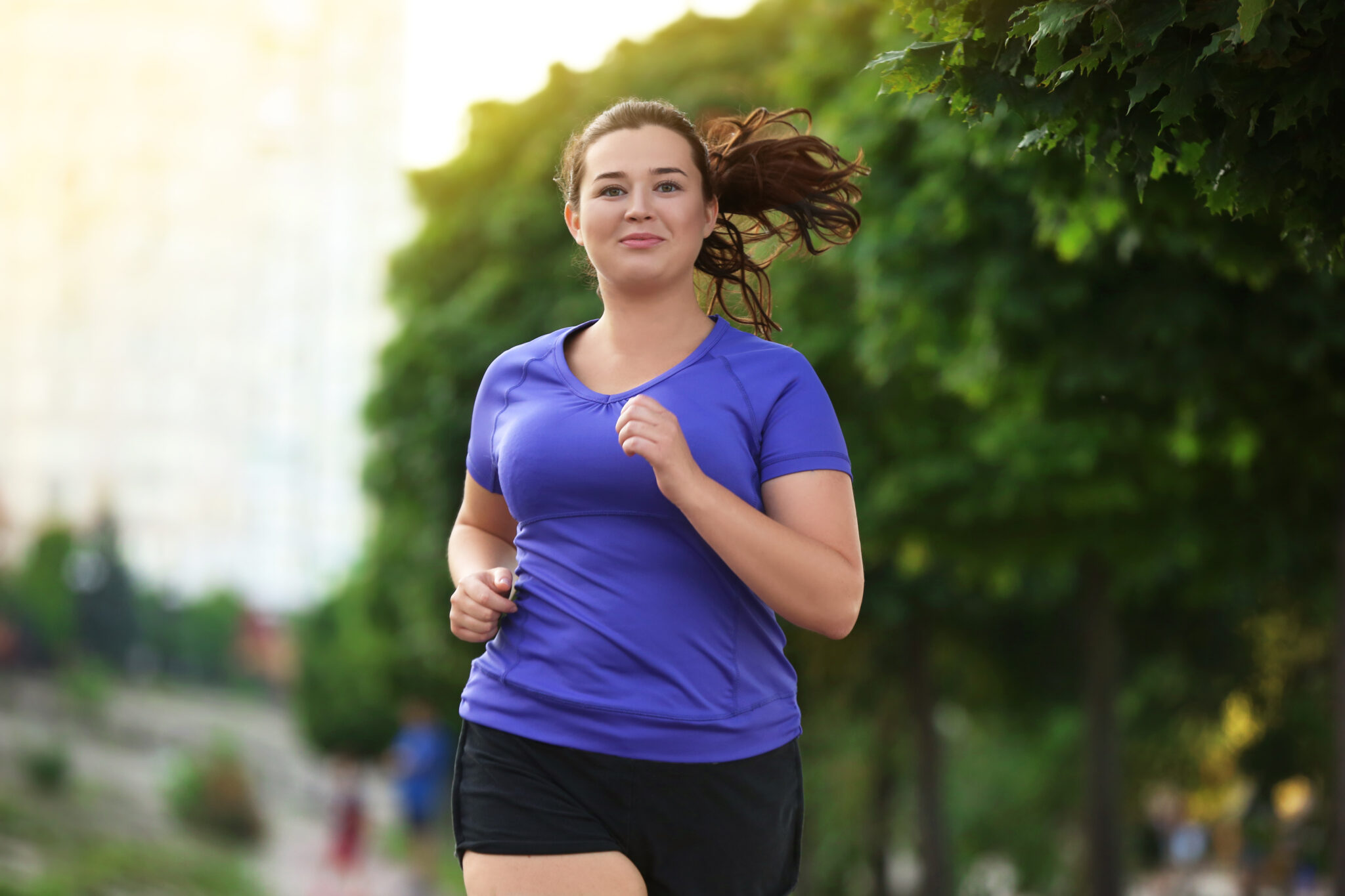 Smiling young woman jogging outdoors in a purple athletic shirt and black shorts, with trees and buildings in the background.