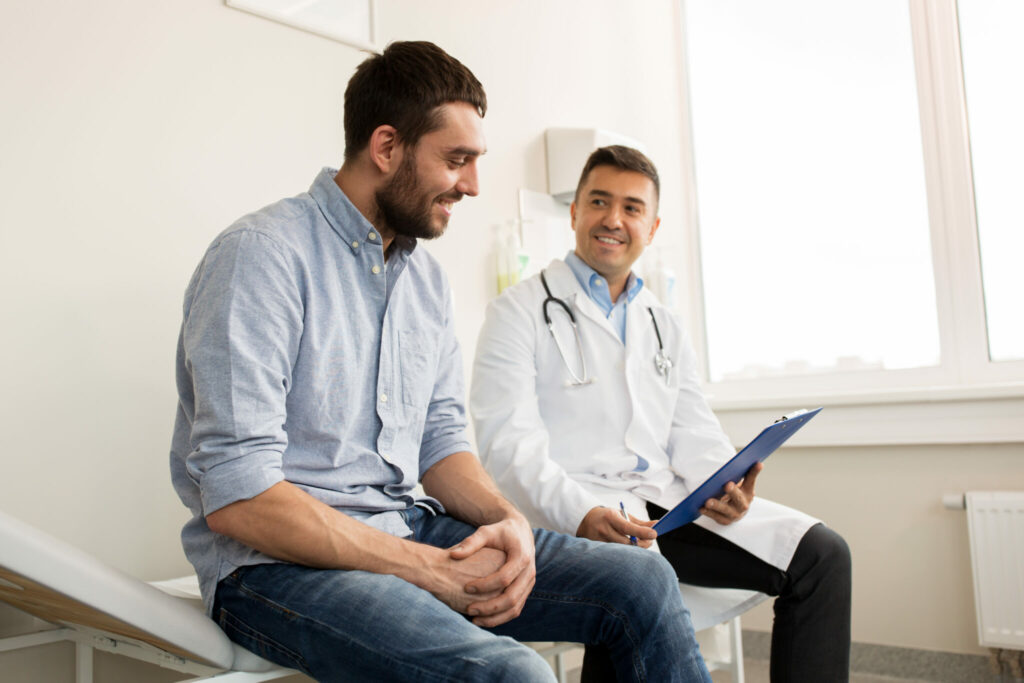 Smiling man in casual clothing sitting on an exam table while a male doctor in a white coat holds a clipboard, both engaged in friendly conversation in a bright medical office.