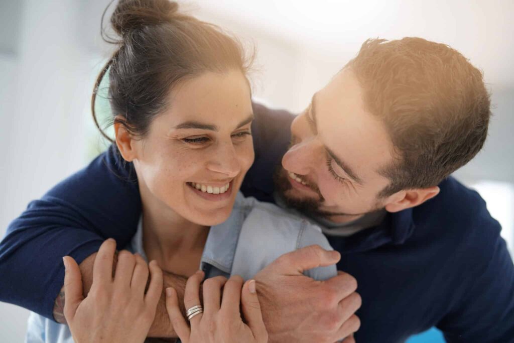 A smiling woman is being embraced from behind by a man, both appearing happy and affectionate in a softly lit, indoor setting.