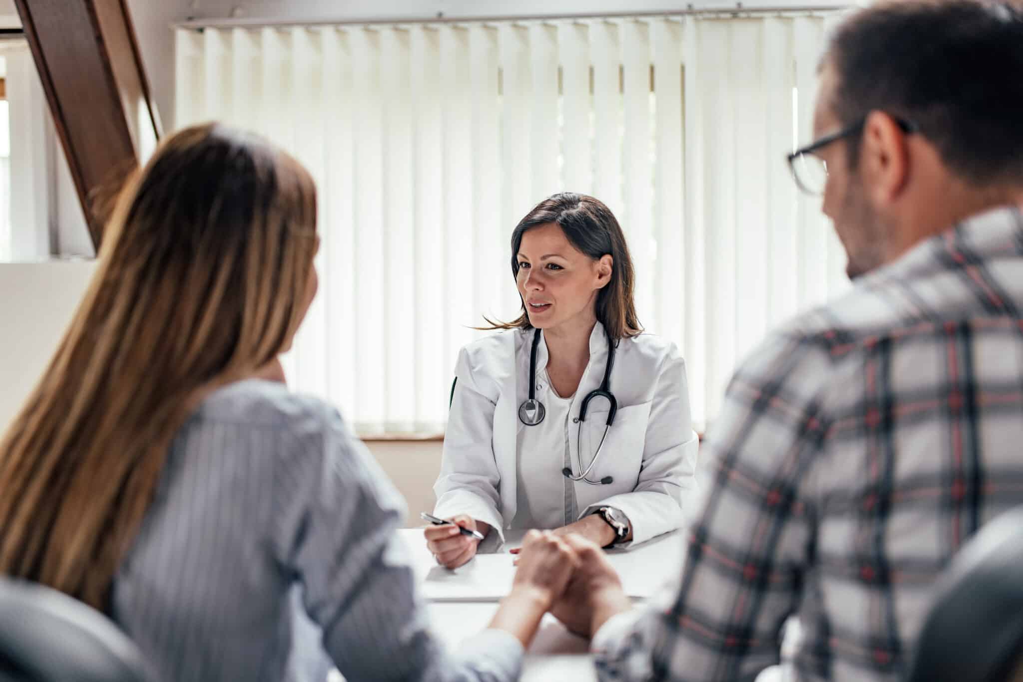 A female doctor is speaking with a couple during a consultation. The doctor is wearing a white coat and a stethoscope, while the couple sits across from her holding hands.