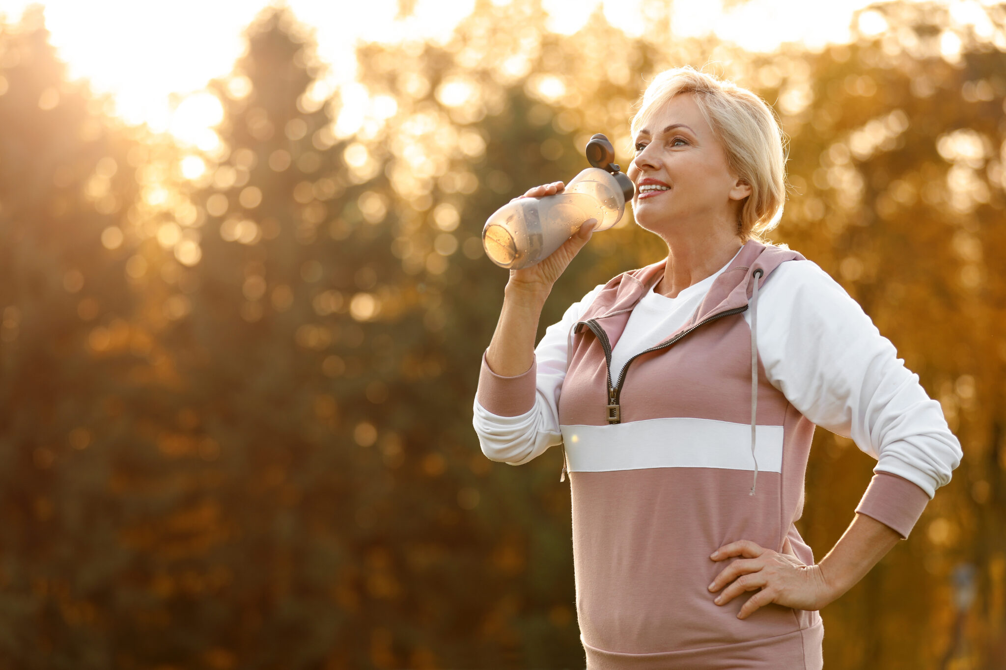 A healthy middle-aged woman drinks water outdoors, wearing workout clothes.