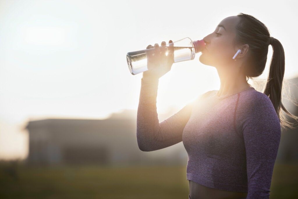 A woman wearing workout attire and wireless earbuds drinks water from a bottle while standing outdoors at sunrise.