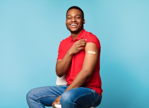 Smiling man in red shirt showing a bandage on his upper arm after receiving an injection.