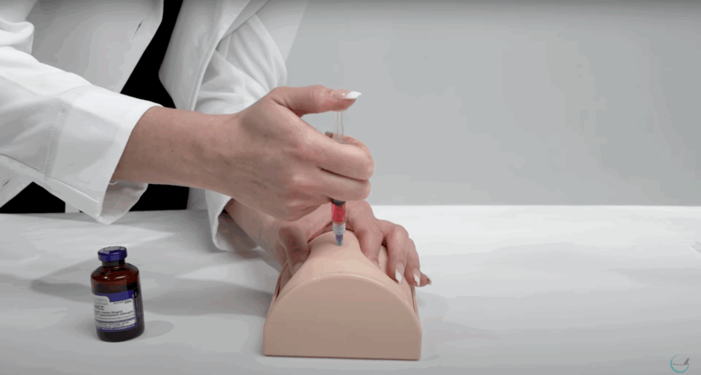 Demonstration of an intramuscular injection using a syringe and a practice pad, with a vial of medication nearby.