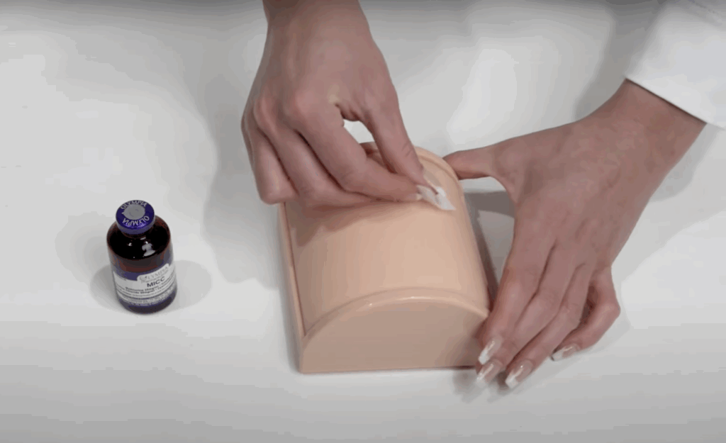 Hands sanitizing a practice injection pad with an alcohol swab next to a vial of Olympia MICC medication.
